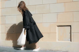 Woman in an oversized black trench coat carrying a white fringed bag along a stone wall