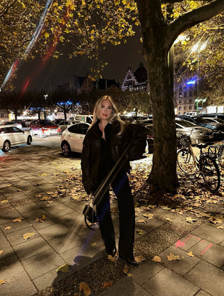 Woman standing on a city street at night with trees and parked cars in the background.