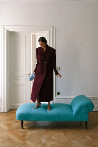 Woman in a long coat standing on a turquoise bench in a room with white walls and wooden floor.